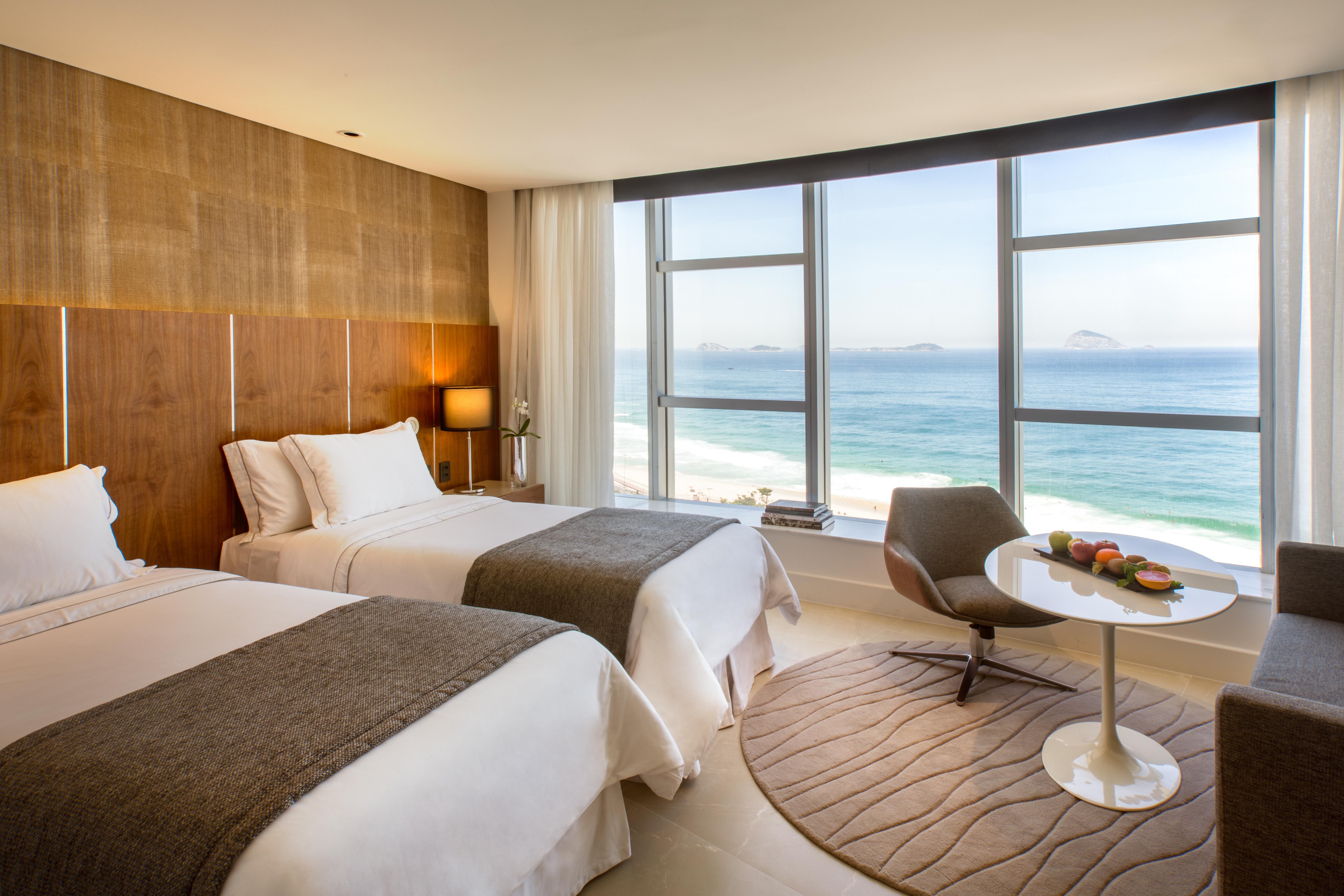 Hotel Nacional Rio De Janeiro - Oficial מראה חיצוני תמונה The photo shows a modern hotel room with two beds adorned in white linens and gray throws. Large windows allow natural light to flood the space, offering a stunning view of the ocean. In the corner, there's a comfortable chair and a small round table